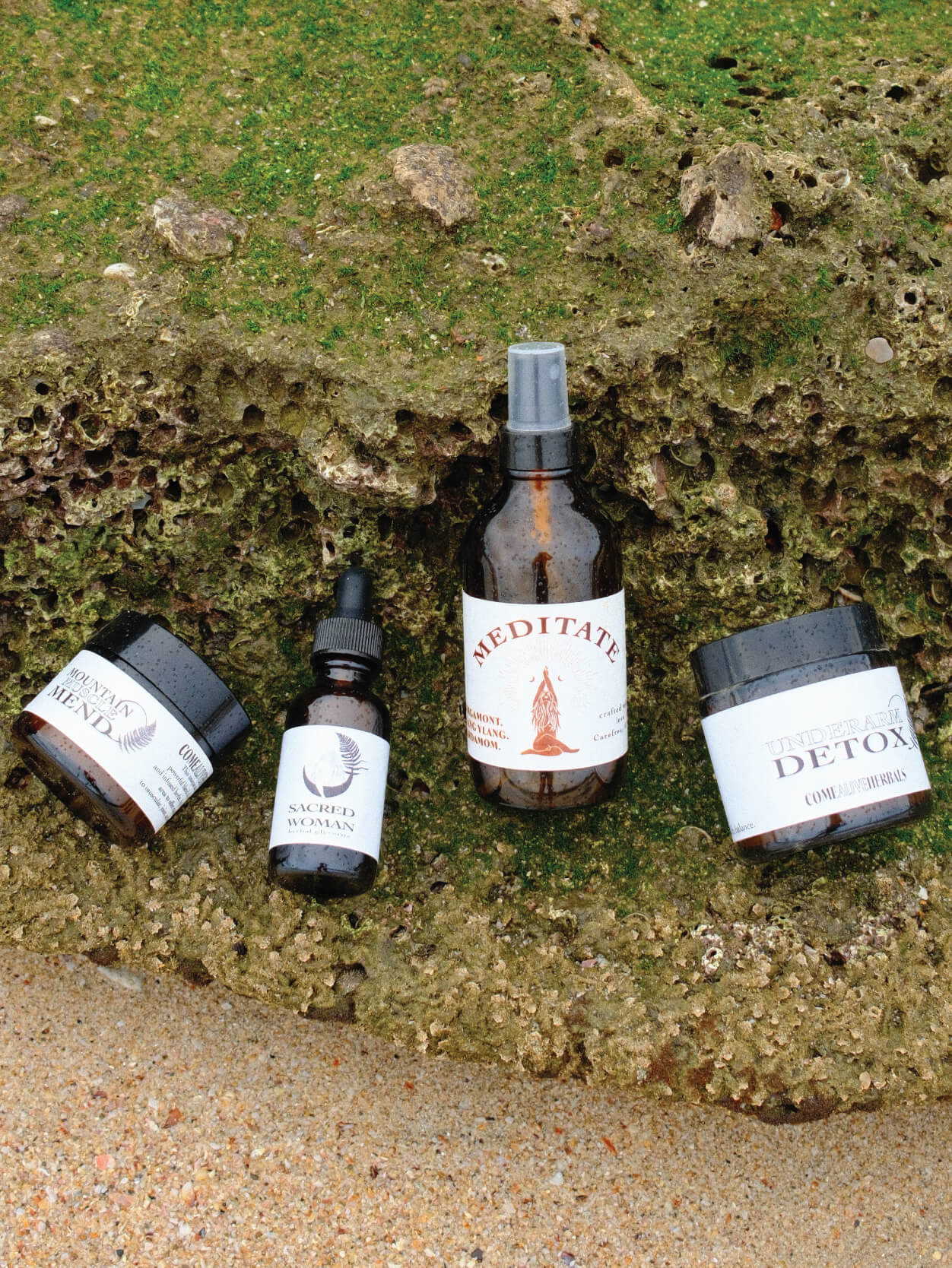 four amber glass come alive herbals products from left to right; mountain muscle mend, sacred woman, meditate mist, underarm detox sitting on a grassy rock surface. 