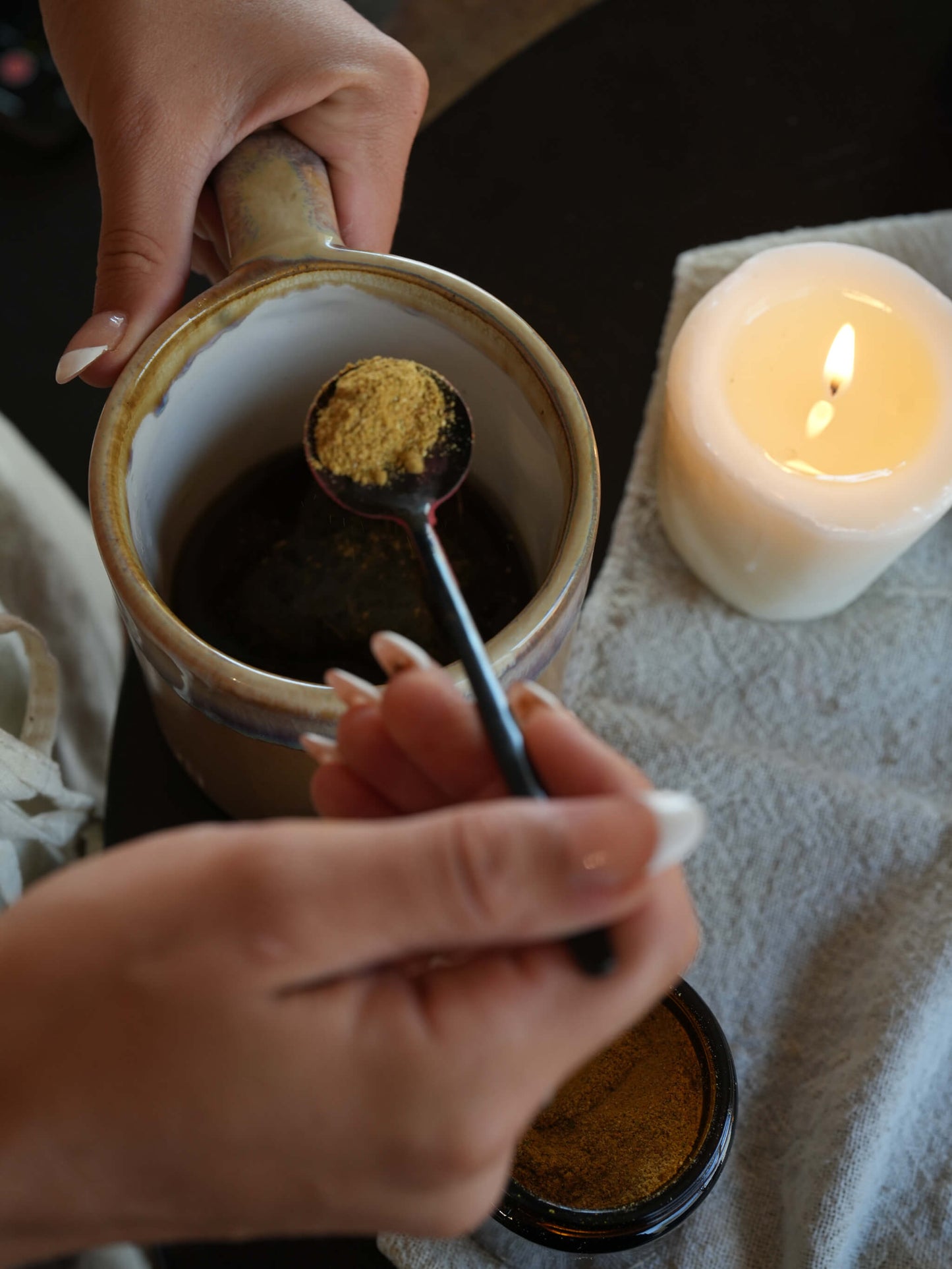 A close-up of hands holding a ceramic mug and spoon while adding a spoonful of golden-brown powder into a dark liquid. A lit white candle sits on a textured towel in the background.