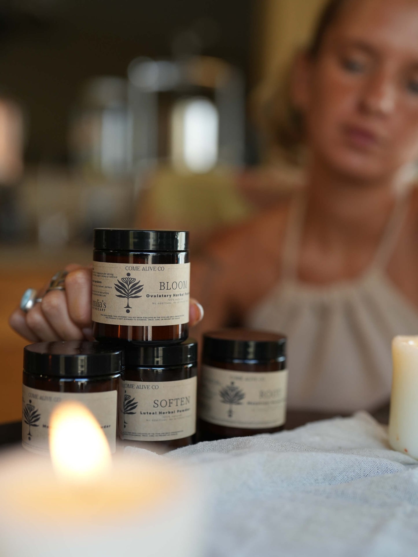 Four amber glass jars of herbal powders with black lids and cream labels are stacked on a table in a spa-like setting, with a woman in a beige top blurred in the background and a lit candle in the foreground.