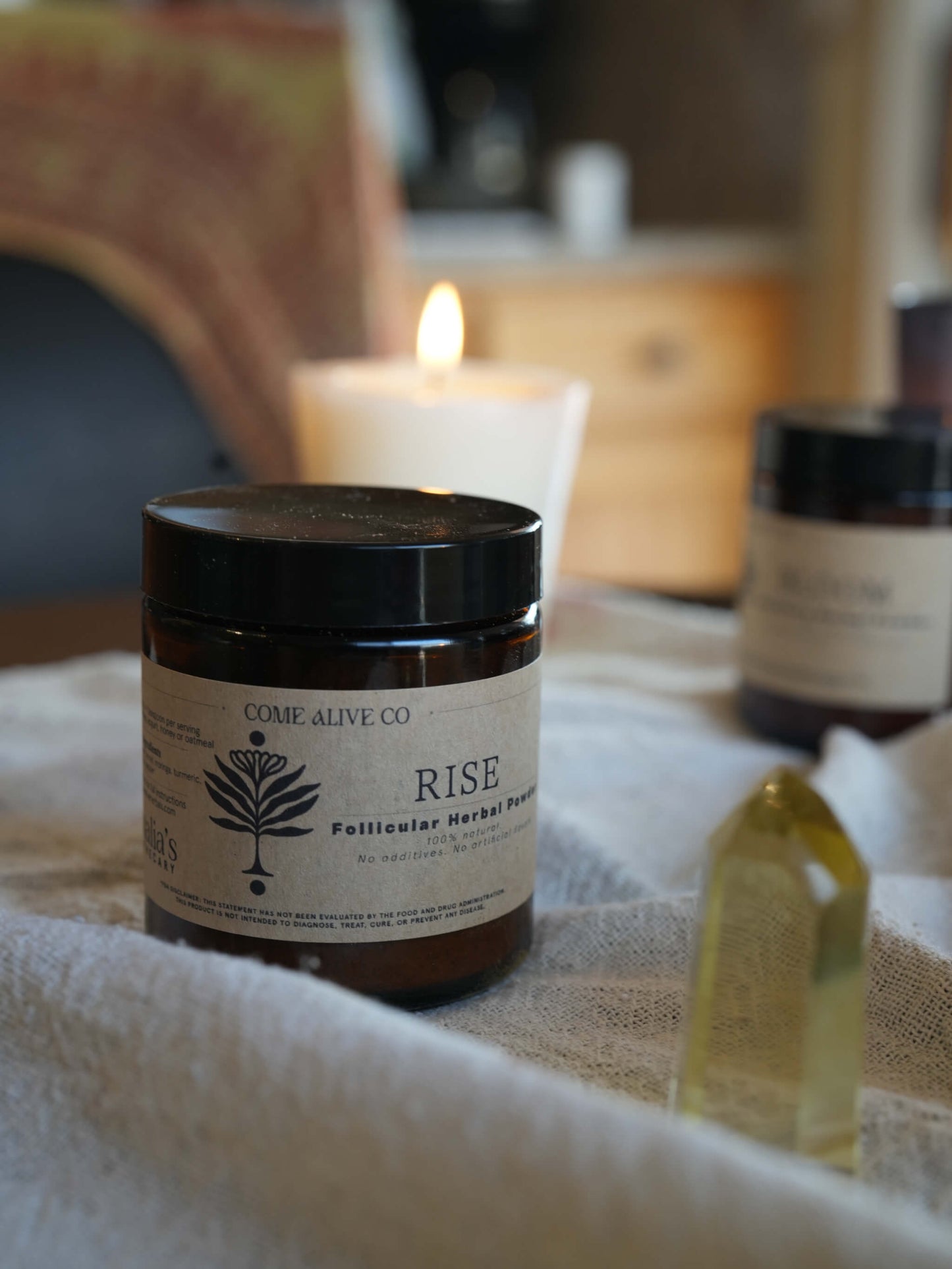 A dark amber glass jar of Come Alive Co. 'Rise' follicular herbal powder sits on a white textured cloth, with a lit candle and other jars blurred in the background.