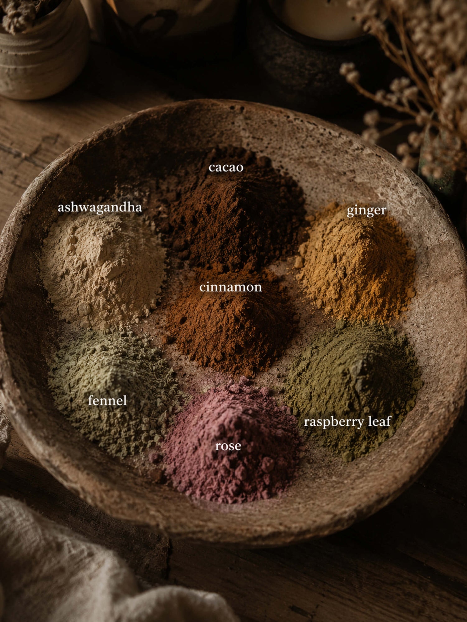 Six loose ground spice blends arranged in small mounds inside a rustic wooden bowl, labeled ashwagandha, cacao, ginger, cinnamon, fennel, and rose. The scene is styled on a wooden surface with dried botanicals and soft warm lighting.