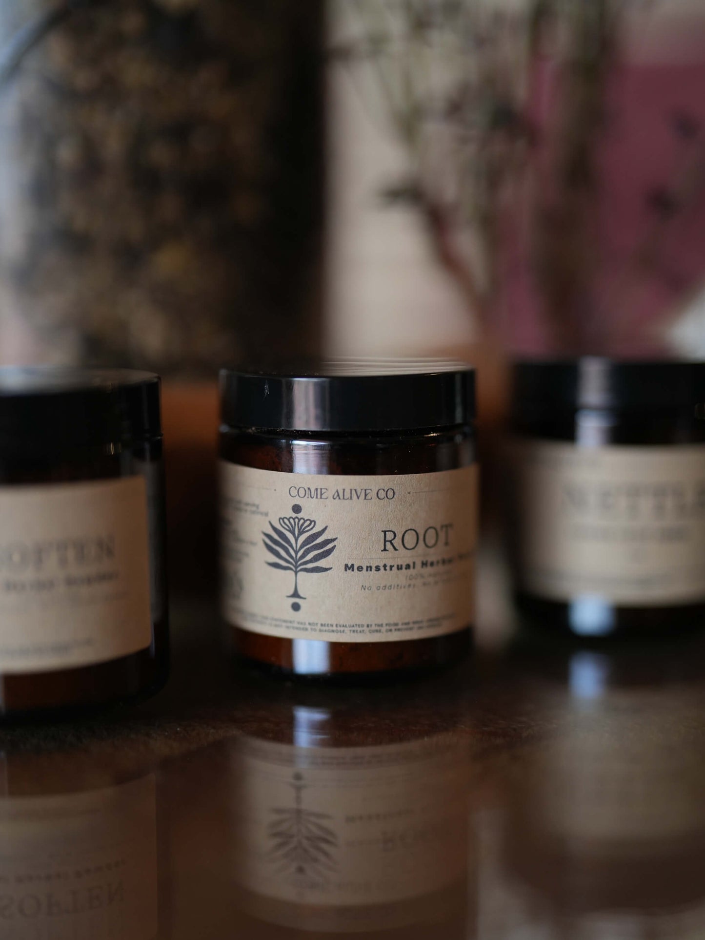 Three amber glass jars with black lids and cream labels are arranged on a reflective dark surface. The center jar is in sharp focus and reads 'COME ALIVE CO.