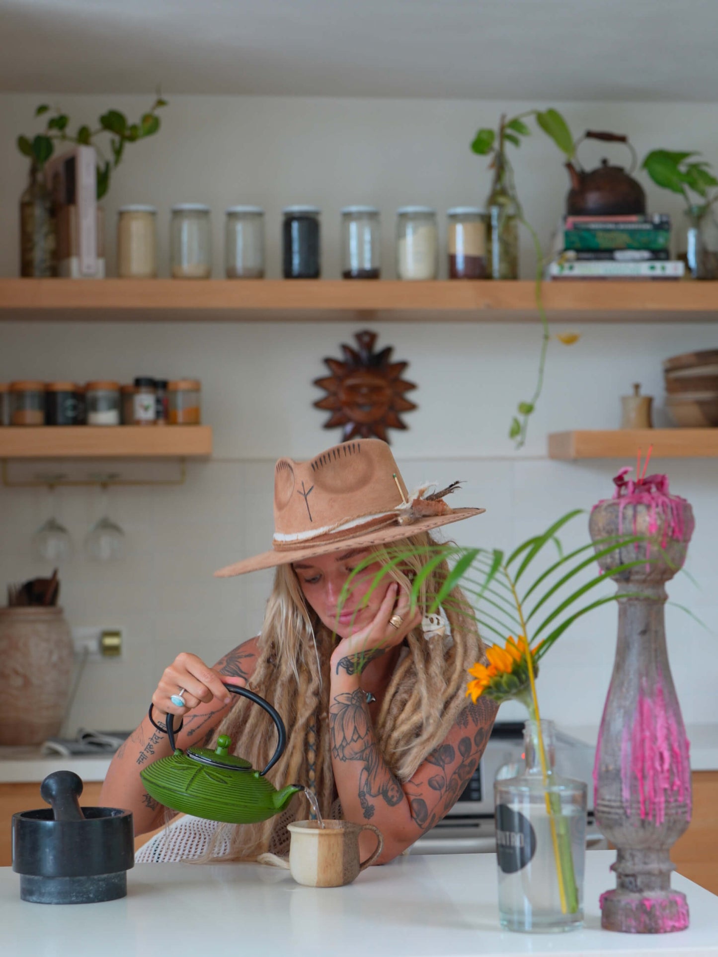 A woman with long blonde dreadlocks sits at a white table in a bright kitchen, pouring water from a green teapot into a small cup. She wears a wide-brimmed tan hat.