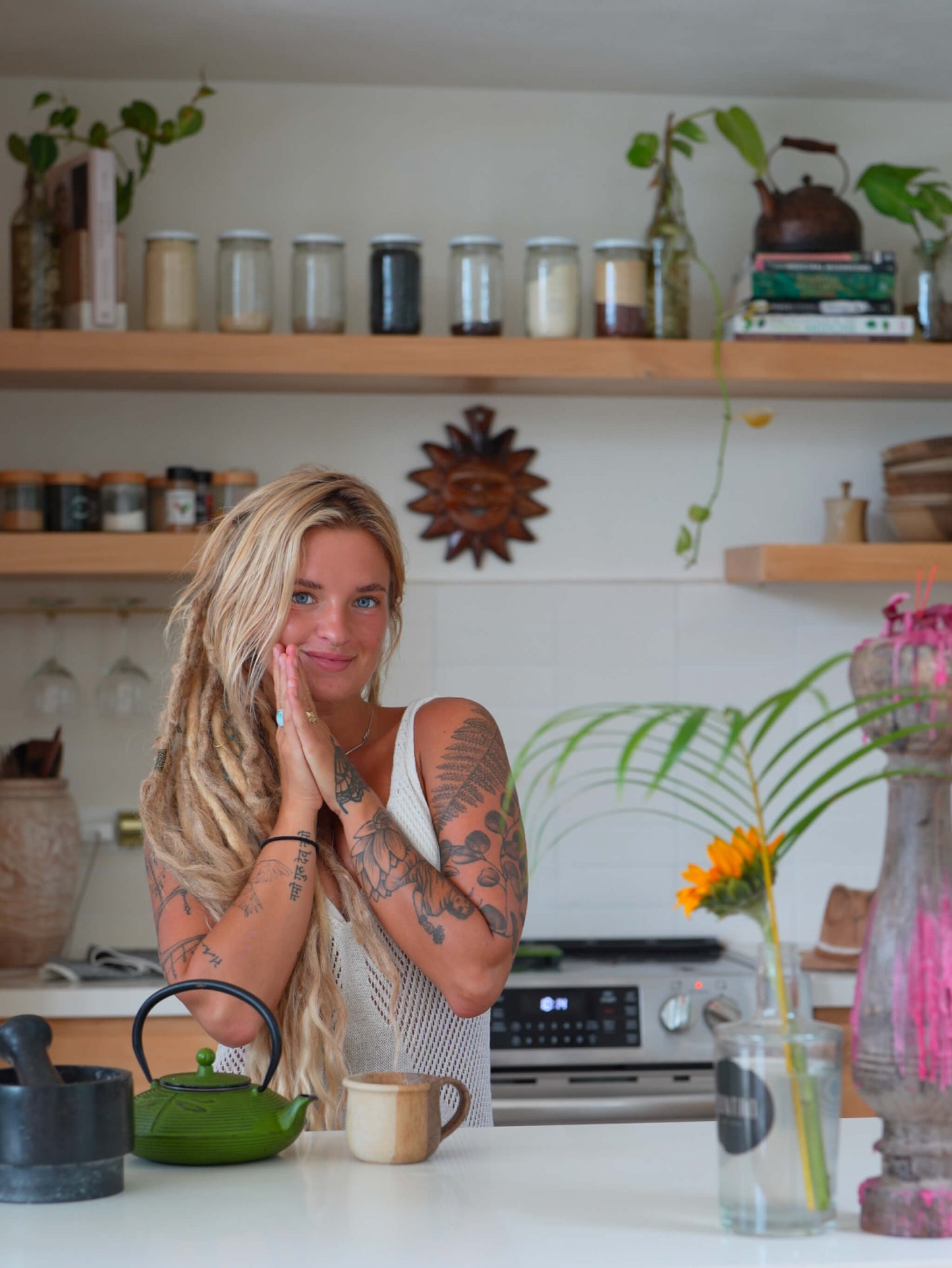 A woman with blonde hair and tattoos sits at a kitchen counter, wearing a white sleeveless top. The background shows a cozy kitchen with wooden shelves, glass jars, and plants.