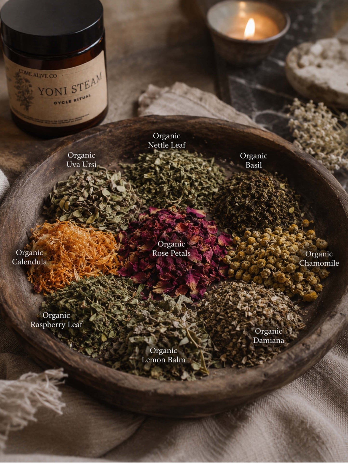 A wooden bowl filled with various dried botanical teas and herbs, arranged in a circular pattern with labels identifying each ingredient. In the background, a jar of Yoni Steam herbal tea and a lit candle are visible on a rustic fabric surface.