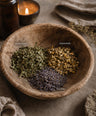 A rustic wooden bowl filled with dried herbal ingredients labeled lemon balm, chamomile, and lavender, styled on a linen surface with a lit candle and amber bottle in the background.
