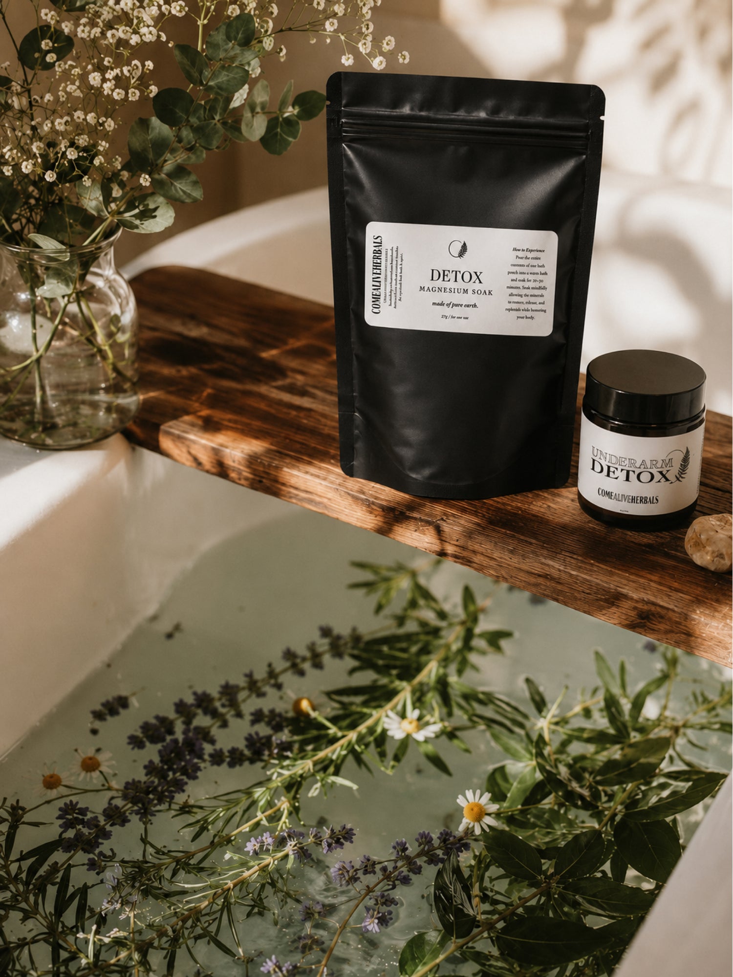 A black stand-up pouch and a small black jar labeled 'Detox' with 'Immunity & Detox' sit on a wooden board beside a bathtub filled with herbs and flowers.