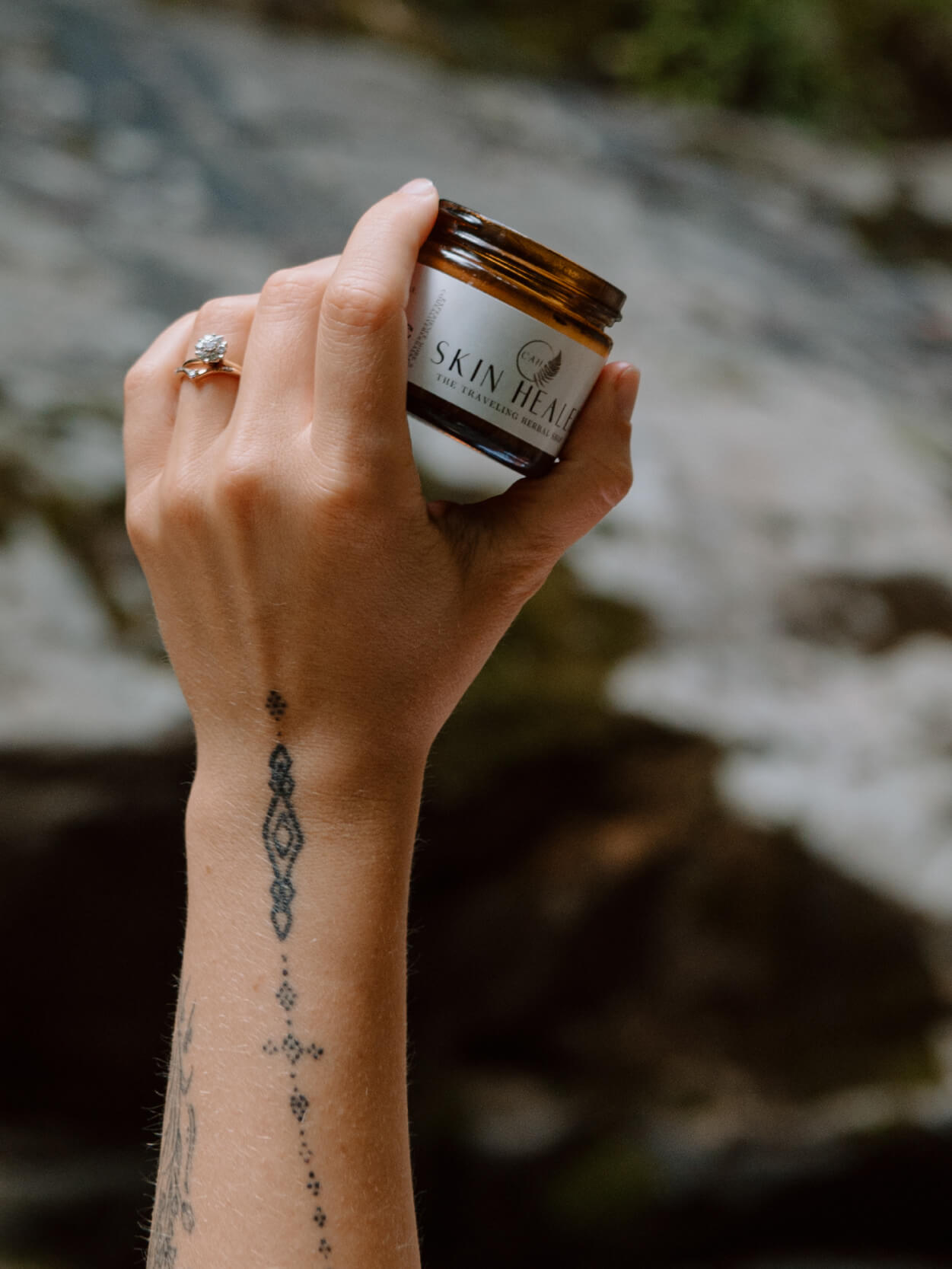 woman hand with tattoo holding amber glass jar of skin healer with label visible with a blurry jungle background. 