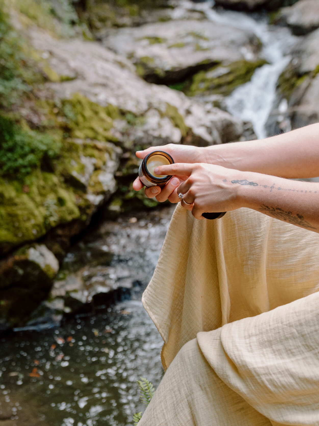 woman hand holding amber glass jar open facing towards picture with other hand dipping finger in jar sitting next to a stream in the jungle wearing beige linen pants. 