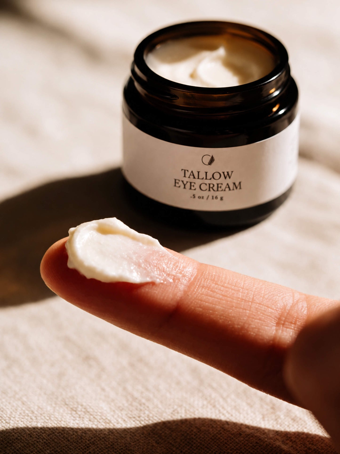 A close-up of a finger holding a dollop of white eye cream in front of an open dark amber jar labeled 'TALLOW EYE CREAM' on a beige fabric background.