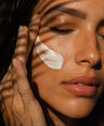 Close-up of a woman's face with closed eyes, applying or showing a white cream swatch on her cheek, with warm sunlight and leaf shadows cast across her face.