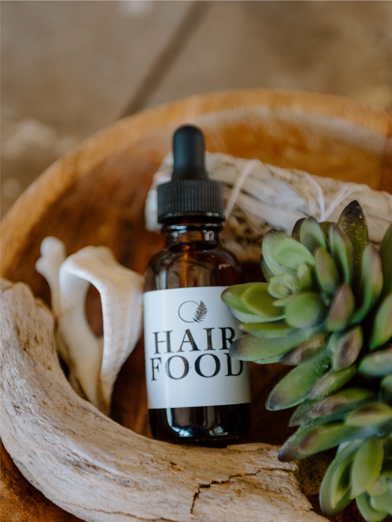 Amber glass bottle with black dropper lid labeled hair food sitting in wooden bowl surrounded by sage bundle, seashell, and succulent plant.