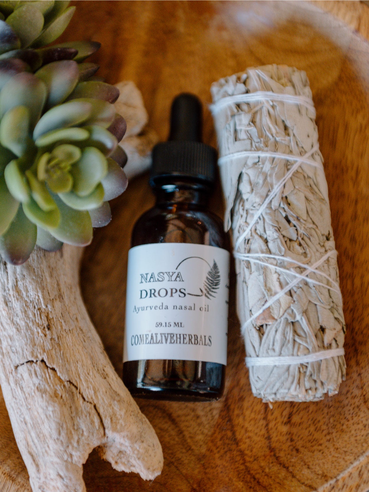 A glass dropper bottle labeled 'Nasya Drops' with a white label and green leaf design, next to a bundle of dried herbs, all placed on a wooden surface. Ayurvedic Nasya Drops for sinus support, breath clarity, and nervous system calm