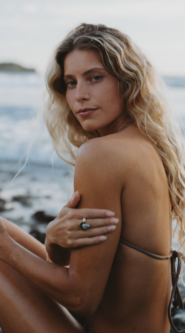 woman rubbing body oil on her arm while sitting on a rock with ocean water in the background