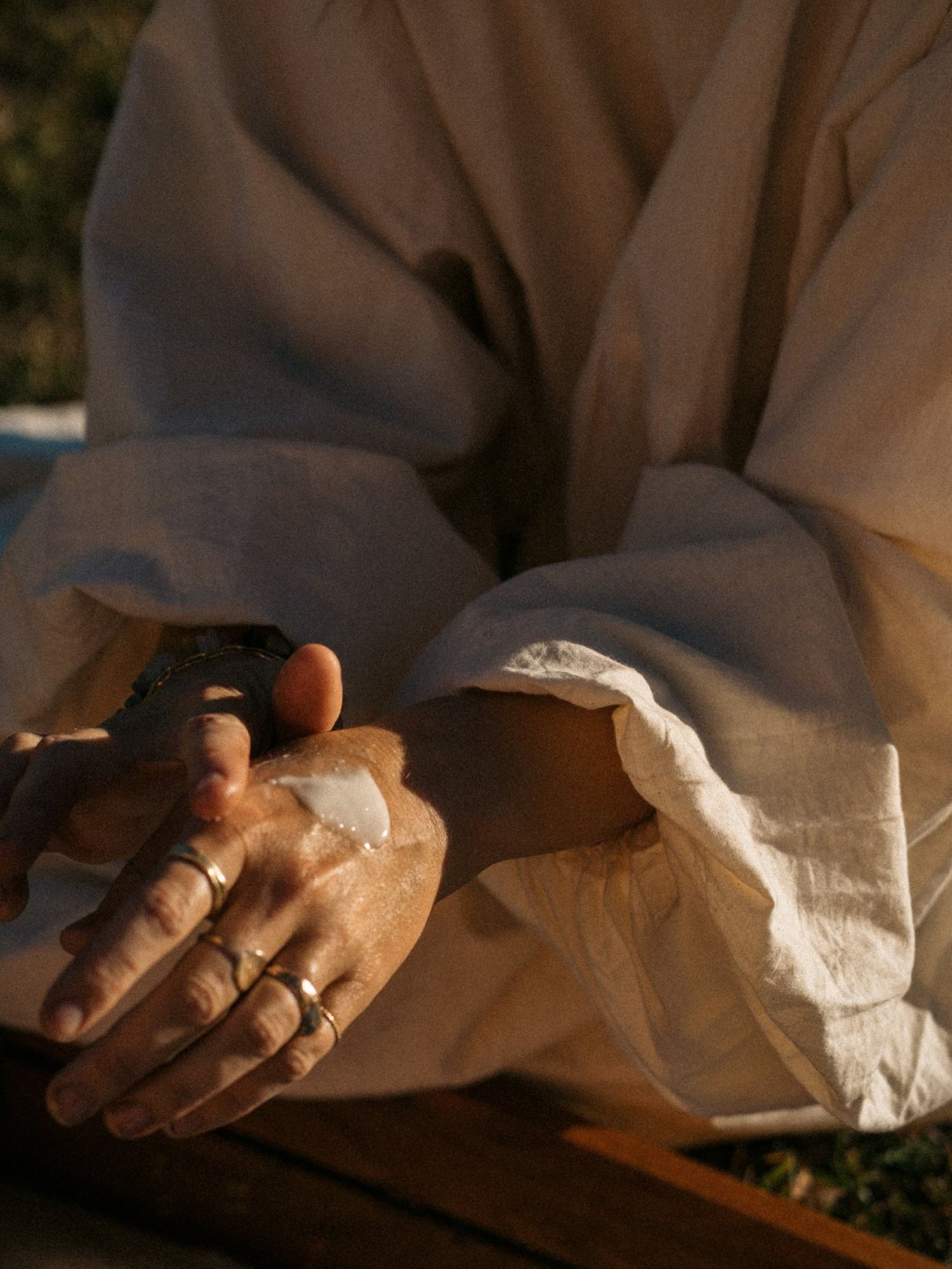 A close-up lifestyle shot of a person wearing a cream-colored linen robe, with their hands clasped together and wearing several gold rings under warm, golden sunlight.