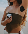 brown haired woman holding amber glass jar labeled underarm detox infront of her chest while other arm is lifted exposing black underarm detox paste on armpit with white background. 