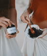one woman hands holding amber glass jar labeled underarm detox while other woman's hands holding seashell with underarm detox paste in shell being mixed by wooden spoon with white background. 