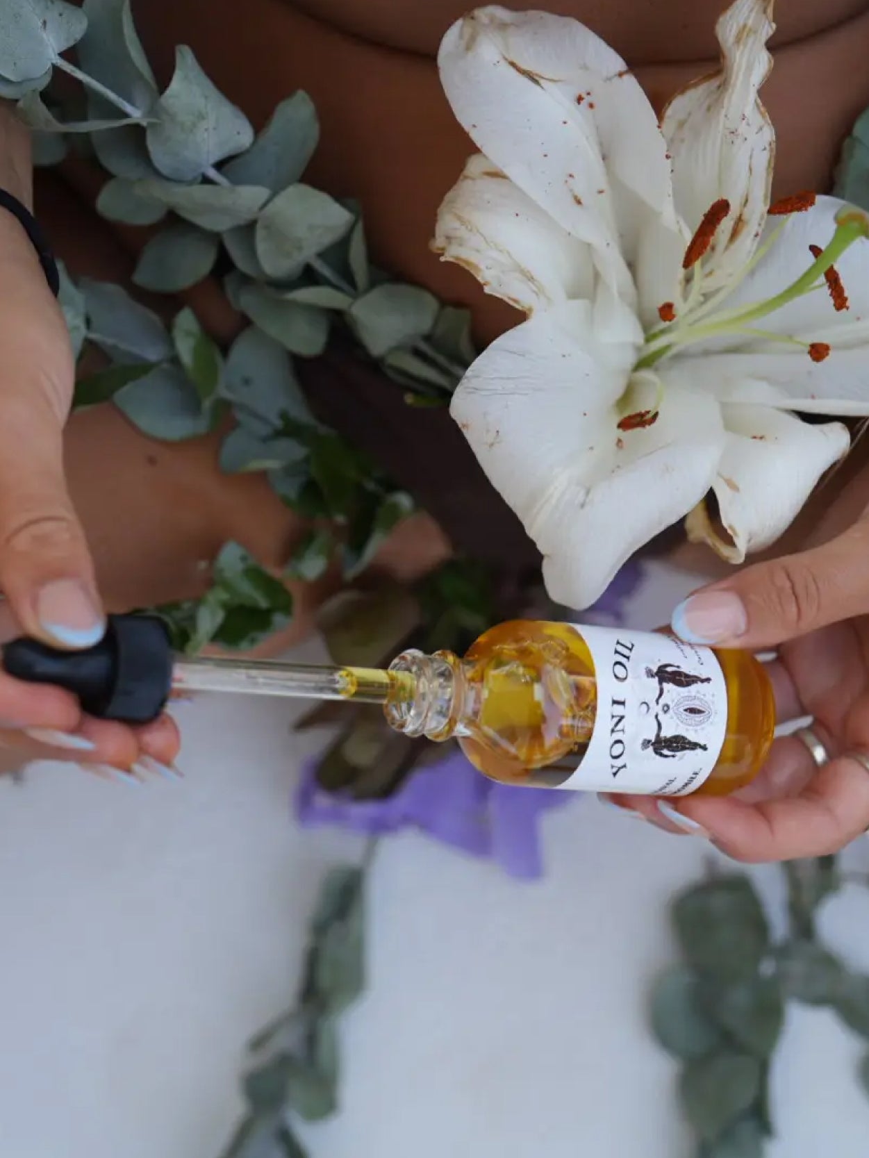 woman hand holding black glass dropper lid with yellow liquid while other hand holds clear glass bottle labeled yoni oil with floral background. 