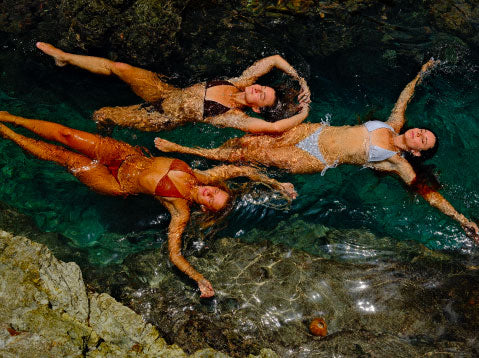 Three women lying on their backs in clear water with rocky surroundings